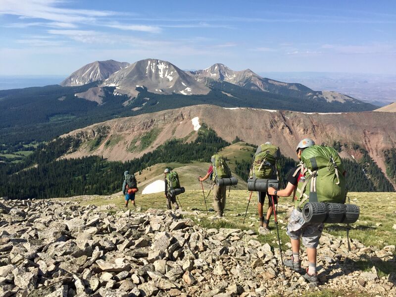 A group of hikers is trekking across a rocky, grassy terrain with large backpacks. In the background, a vast mountain range stretches under a clear blue sky. The hikers are spread out, suggesting a challenging but scenic hike. The landscape is a mix of green vegetation and exposed rock, typical of a high-altitude environment.